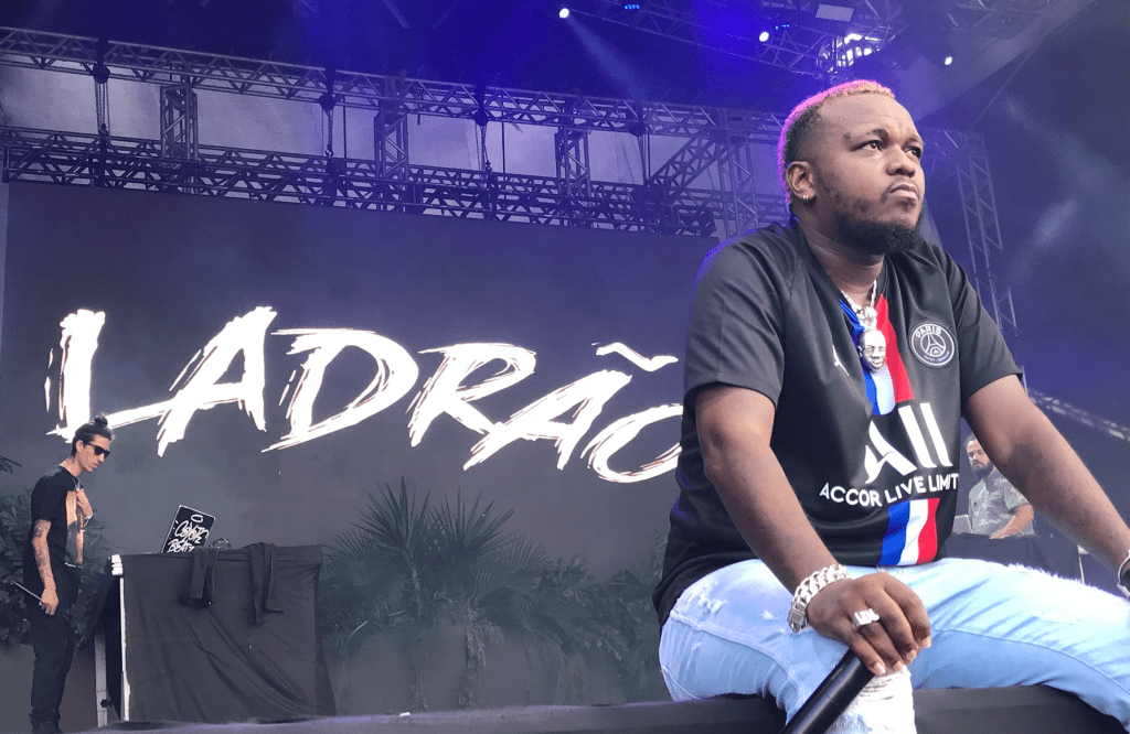 Rapper Djonga sentado sobre o palco e olhando para o horizonte. Ele é um homem preto, está com a camisa escura do time de futebol PSG e veste uma calça jeans clara. Ao fundo, no cenário do palco, está escrito LADRÃO