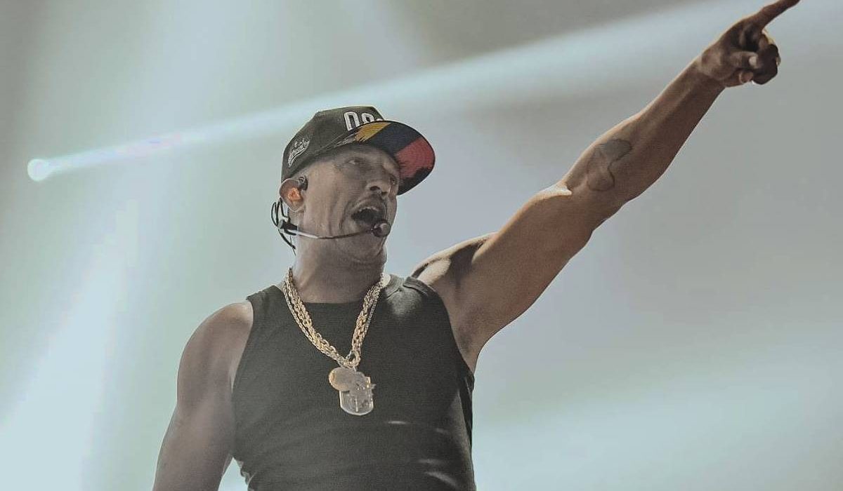 Mano Brown está no palco com uma camiseta regata preta, cordão de ouro e boné preto. Ele levanta o braço esquerdo e aponta para a frente