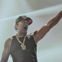 Mano Brown está no palco com uma camiseta regata preta, cordão de ouro e boné preto. Ele levanta o braço esquerdo e aponta para a frente