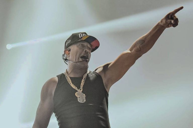 Mano Brown está no palco com uma camiseta regata preta, cordão de ouro e boné preto. Ele levanta o braço esquerdo e aponta para a frente